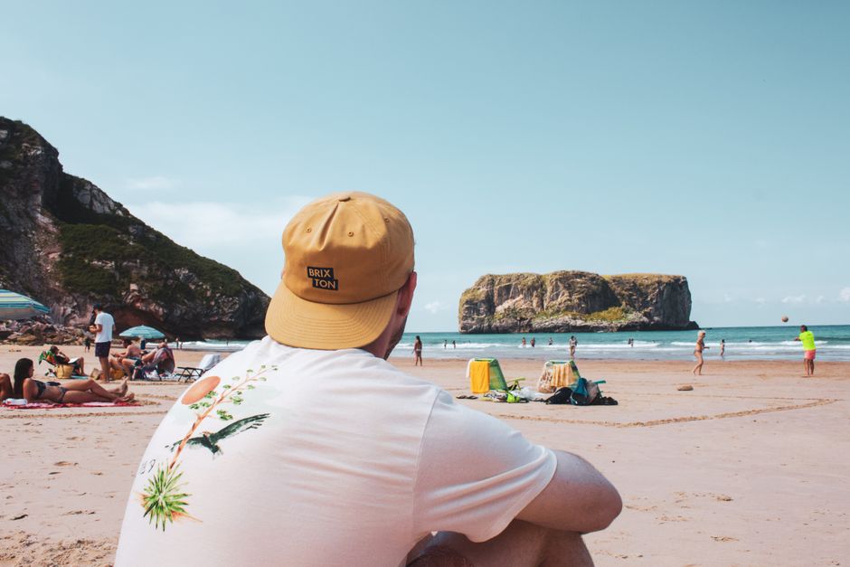 A man wearing a beige bucket hat with a 'BRIXTON' label and a white t-shirt featuring a tropical bird and plant illustration sits on a sandy beach facing the sea, with his back to the camera. In the background, there are several beachgoers sunbathing, walking along the shoreline, and relaxing under umbrellas, with their belongings such as bags and towels scattered on the sand. The coastline is framed by green, rocky cliffs on the left and a large, flat-topped sea stack on the right, with calm ocean waves gently breaking near the shore. The scene is illuminated by natural daylight, suggesting a clear, sunny day. Although not directly depicted, this setting reflects the importance of planning for outdoor activities and parking during a house move or home relocation in Brixton, as highlighted in the 'Acre Lane removals and parking tips for Brixton SW9' page on Brixton Man and Van's website, which provides guidance on efficient moving logistics.