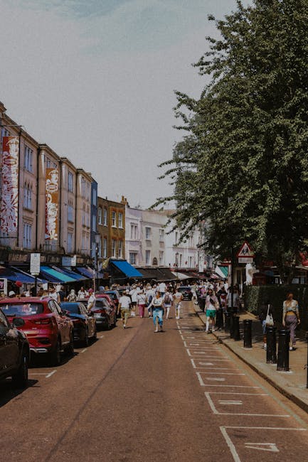 The image depicts a lively urban street scene in Brixton with a large crowd of pedestrians walking along the pavement and across the road. On the left side of the image, rows of parked cars line the street, including various models and colours. The right side features large trees providing partial shade, and bollards separating the pedestrian walkway from the road. In the background, multi-storey buildings with a mix of retail shops, cafes, and bars have awnings and signage, indicating commercial activity typical of a busy shopping area. The scene is captured during daytime with clear skies, and the lighting highlights the vibrant atmosphere. The context relates to house removals and moving services, suggesting the importance of parking management and logistical planning in urban areas like Brixton, where [COMPANY_NAME] offers professional removals and parking assistance for home relocations in SW9 neighborhoods, ensuring smooth and efficient furniture transport during home moves.