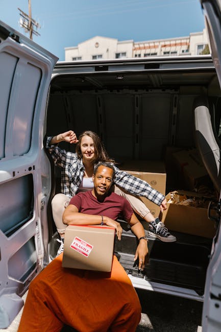Two young adults, a woman and a man, are sitting on a staircase inside a building, both smiling and holding cardboard boxes typically used for home relocation. The woman, with long hair, is seated on 