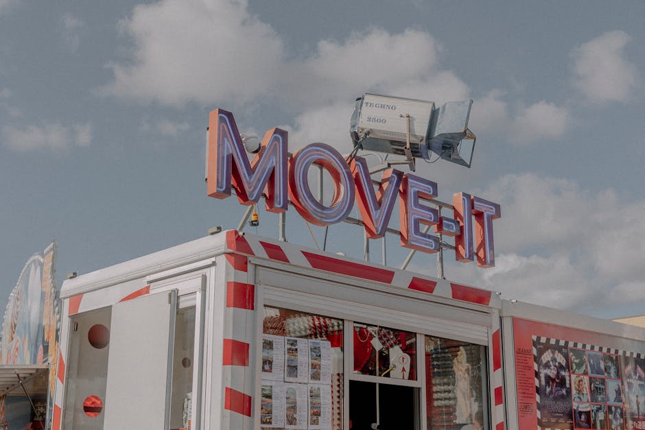 A large mobile unit with a white exterior featuring red reflective stripes, situated outdoors on a paved surface under a partly cloudy sky. The unit has a prominent illuminated sign on top spelling 'MOVE-IT' in bold, multicoloured lights, with a professional lighting fixture positioned above the sign. The structure appears to be part of a house removal or furniture transport setup, potentially used for packing, storing, or staging items before a move. Visible on the side of the unit are various posters and advertisements related to moving services, along with clear windows revealing interior shelves and packed boxes. Nearby, there are additional items such as a cardboard box and moving blankets, indicating preparation for home relocation tasks. The scene reflects a professional moving process, with Brixton Man and Van providing a mobile solution for furniture transport and packing in the context of house removals or local moves happening in the Brixton area.