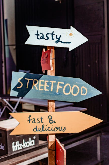 A multi-directional wooden signpost with four colorful arrow-shaped signs is positioned outdoors against a dark wall. The top sign, painted white, reads 'tasty' with small orange accents. Below it, a blue sign points to 'STREET FOOD' in large beige letters. The third sign, painted yellow-orange, states 'fast & delicious' in handwritten style. The signs are mounted on a vertical wooden post, which is anchored into the ground. In the background, part of a window and a bench with a Fitz-Kola soda crate are visible, indicating a casual outdoor setting that may be near a marketplace or food area. This setting is captured in natural light, highlighting the vibrant colors and clear text on each sign, and is relevant to the context of city living and movement, echoing themes of packing, carrying, or transporting goods during house removals or urban relocations, as handled by Brixton Man and Van.
