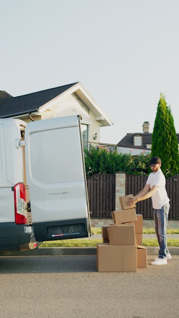 A man stands outdoors beside a white moving van with its side door open, preparing to load or unload cardboard boxes of various sizes situated on the pavement. The boxes are sealed with packing tape, some are stacked on top of each other, and appear to contain household items for a home relocation. The man is dressed in casual clothing, wearing a white t-shirt and dark jeans, and is holding a cardboard box. Behind him, there is a residential area with a garden featuring a trimmed hedge, a tall coniferous tree, and a cream-colored house with a dark roof. The scene is well-lit with natural daylight, indicating a clear day, and the surroundings suggest a typical urban or suburban street, with no other vehicles or people visible. This visual supports the process of furniture transport and packing as part of house removals, with the involvement of Brixton Man and Van in standard moving services for tight access locations, as indicated by the page title for a detailed guide on house removals on Coldharbour Lane. 