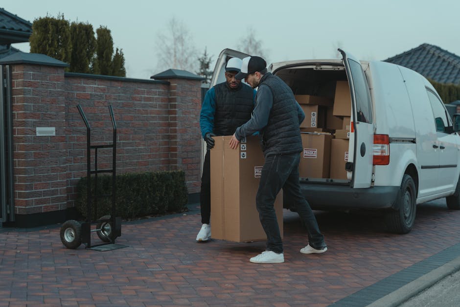 Two movers from Brixton Man and Van are inside a driveway loading a large cardboard box into the rear of a white panel van, which is parked near a brick wall and residential houses. One worker is holding the box steady while the other guides it into the vehicle. A black hand truck with two wheels is standing nearby on the brick-paved surface. The van’s rear doors are open, revealing additional boxes stacked inside, some made of cardboard and secured with plastic wrap. The scene is set outdoors during overcast weather, with trees and rooftops visible in the background, illustrating a professional home relocation or furniture transport process involving packing and loading equipment.