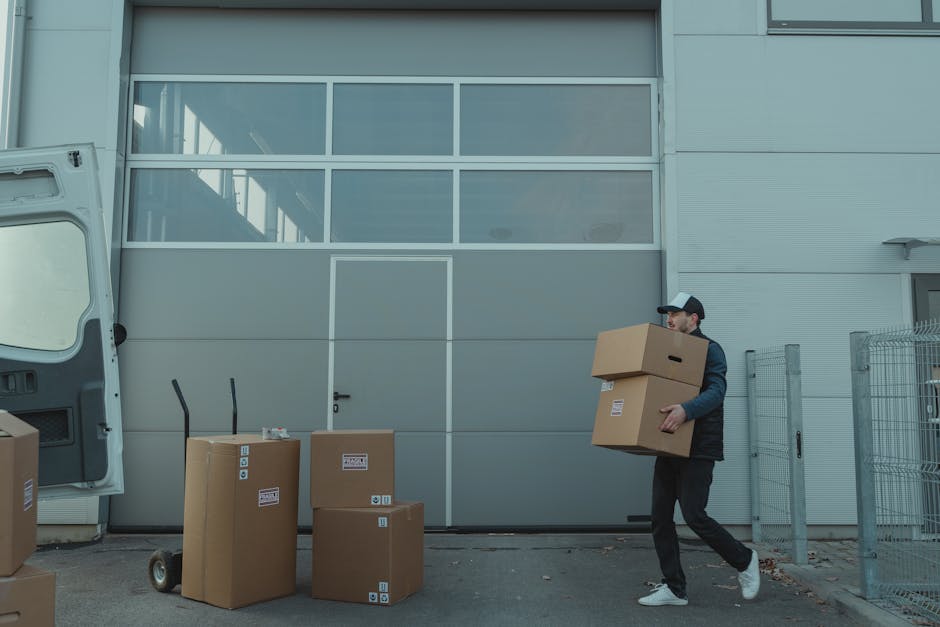 A man stands outdoors beside a white moving van with its side door open, preparing to load or unload cardboard boxes of various sizes situated on the pavement. The boxes are sealed with packing tape, some are stacked on top of each other, and appear to contain household items for a home relocation. The man is dressed in casual clothing, wearing a white t-shirt and dark jeans, and is holding a cardboard box. Behind him, there is a residential area with a garden featuring a trimmed hedge, a tall coniferous tree, and a cream-colored house with a dark roof. The scene is well-lit with natural daylight, indicating a clear day, and the surroundings suggest a typical urban or suburban street, with no other vehicles or people visible. This visual supports the process of furniture transport and packing as part of house removals, with the involvement of Brixton Man and Van in standard moving services for tight access locations, as indicated by the page title for a detailed guide on house removals on Coldharbour Lane. 